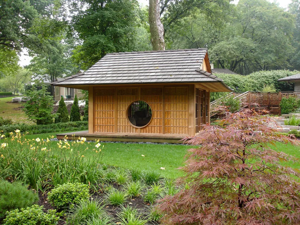 Wood garden pavilion bordered by healthy lawn, layered perennial planting, shrubs, and mature trees